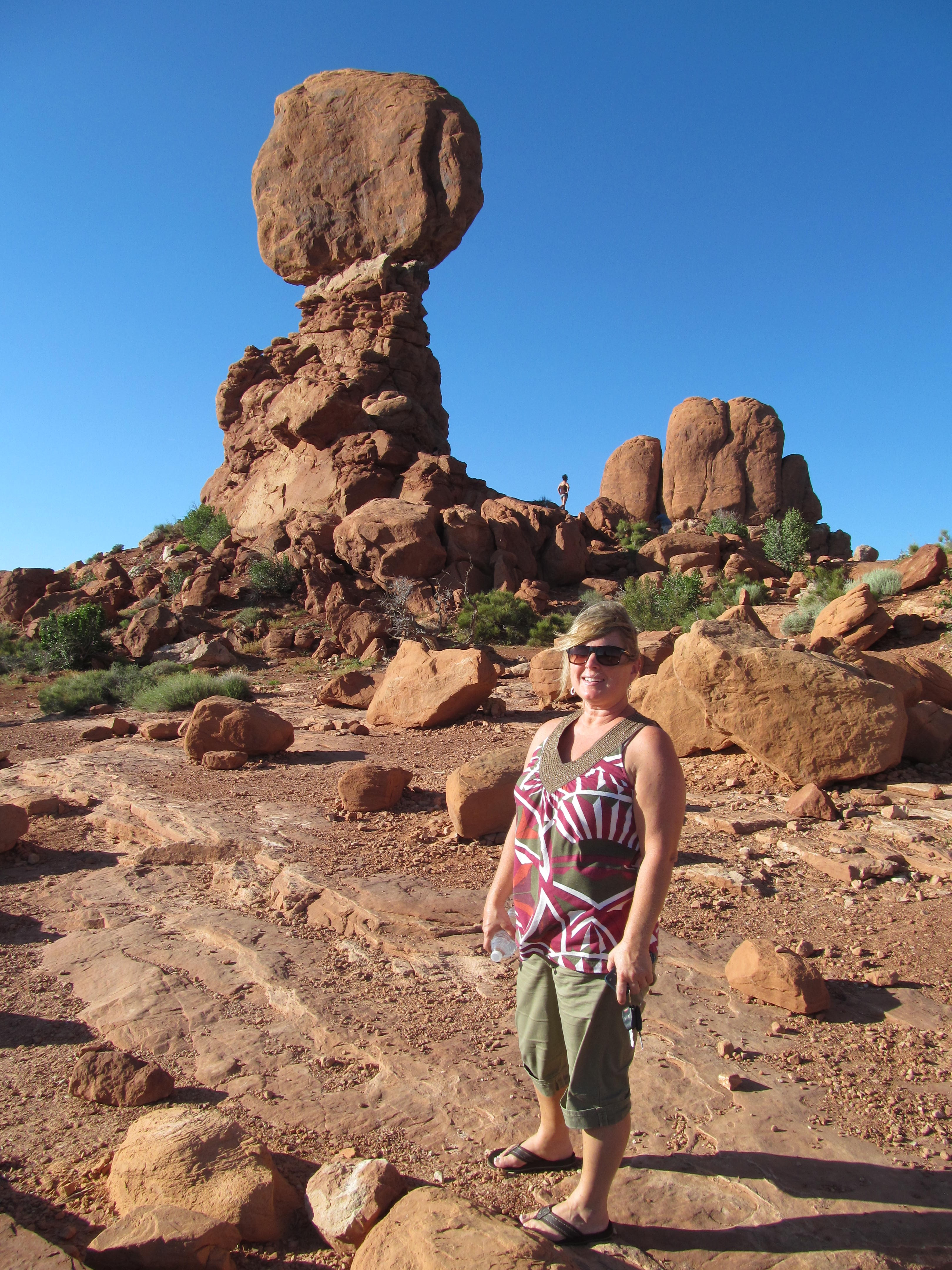 Cindy standing below Balanced Rock beneath a perfectly clear desert sky.