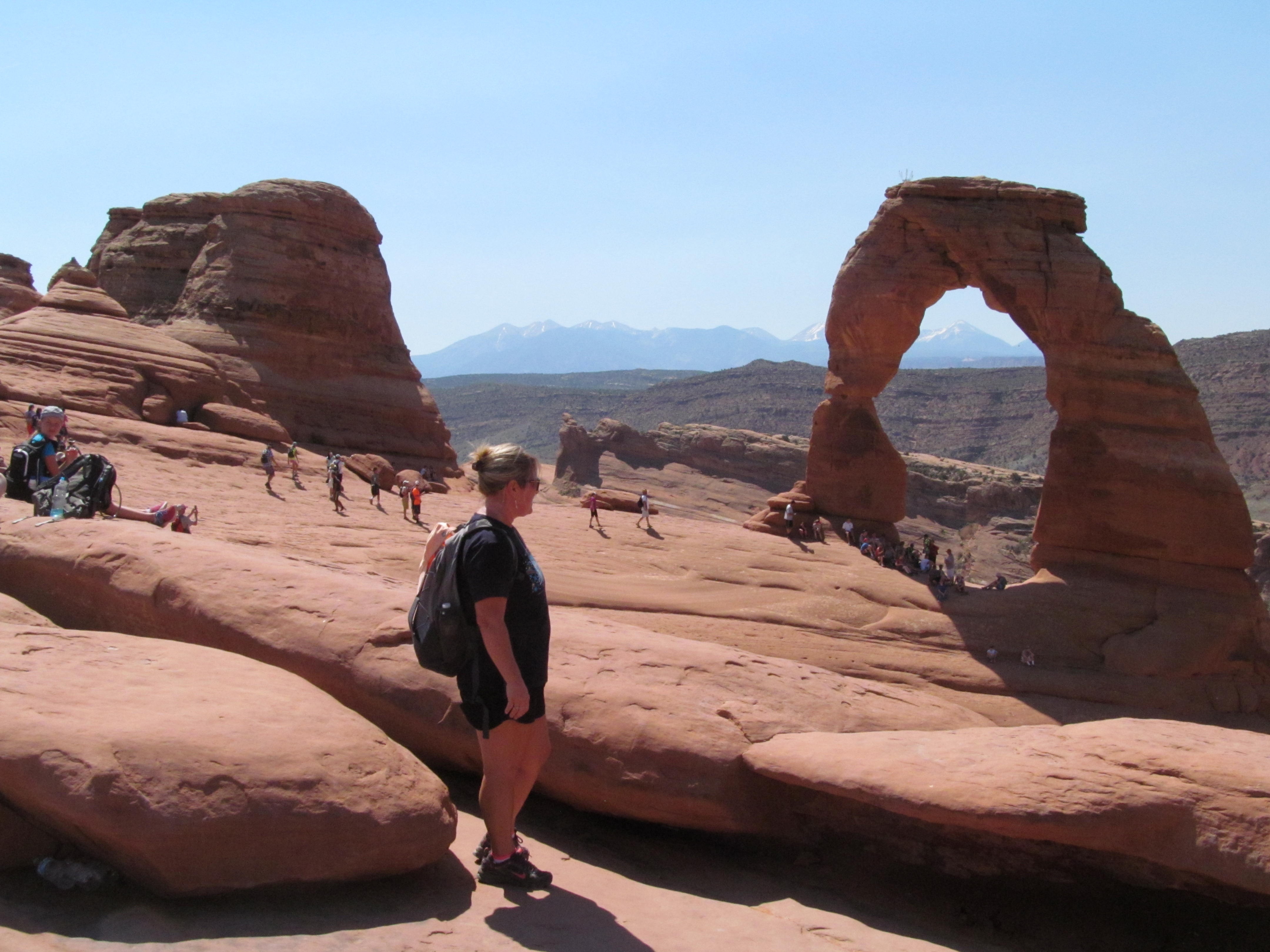 Cindy with Delicate Arch standing above the sweeping sandstone bowl.