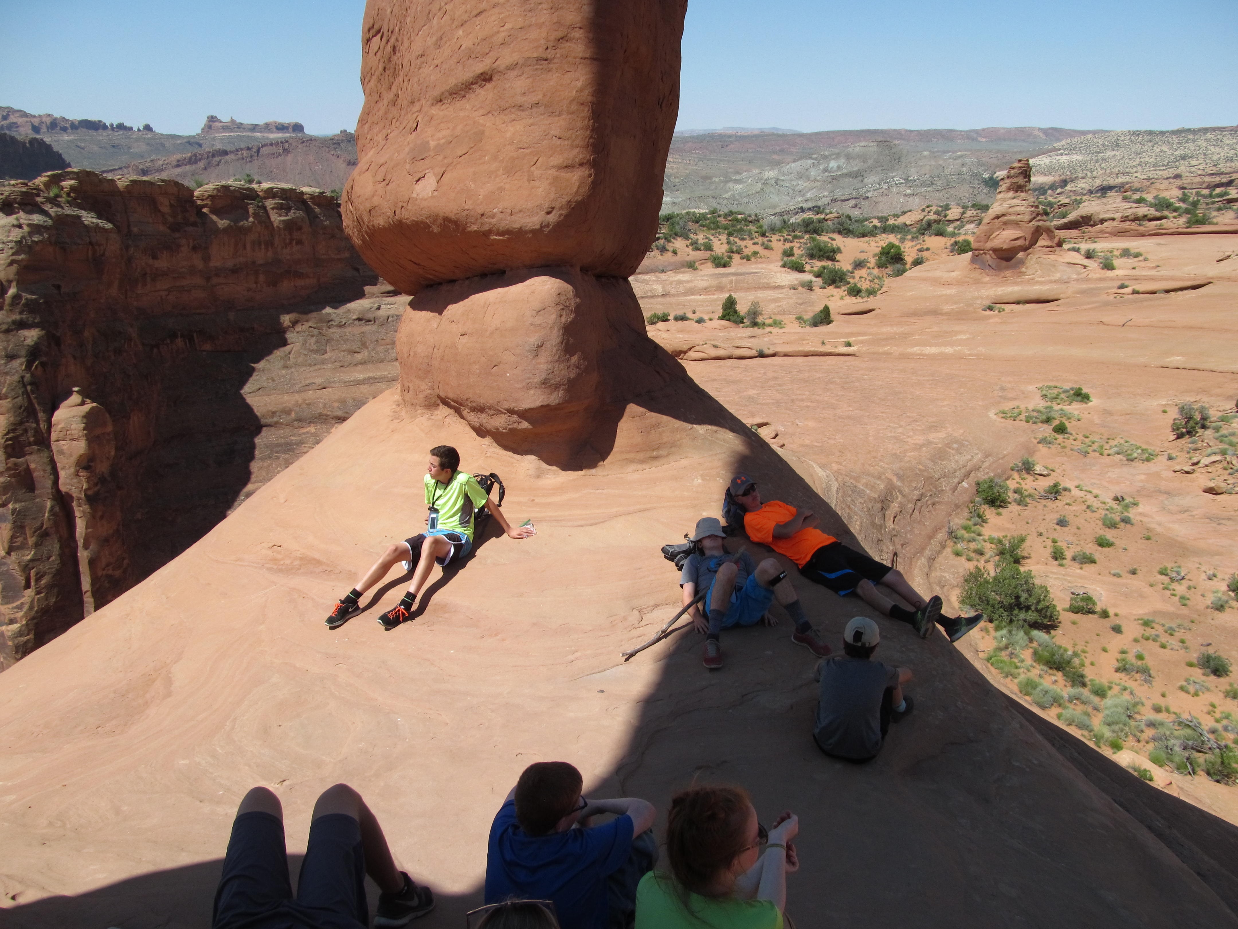 Hikers resting in a patch of shade beneath a sandstone wall.