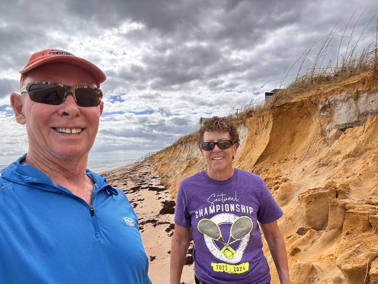 Tim and Kym explore a beach backed by dramatically eroded sand bluffs beneath stormy skies.